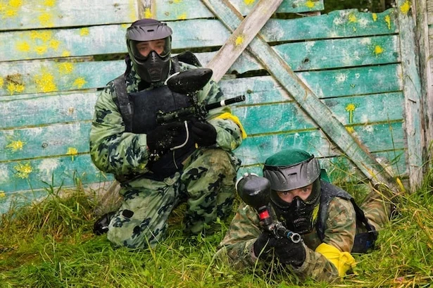 Dos jugadores con caretas y equipo de paintball junto a una barrera de madera, en una foto publicada por La Reserva